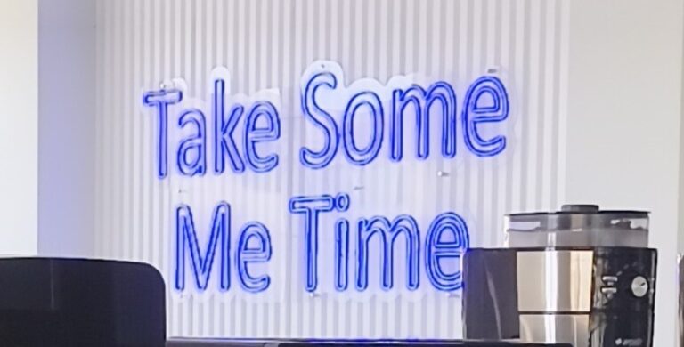 A neon sign reading 'Take Some Me Time' above a coffee machine